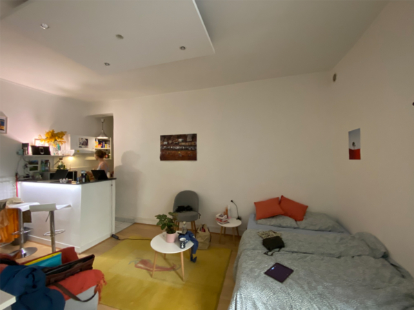 Interior view of a compact studio apartment showing a person at a kitchen counter, a bed with orange pillows, a small coffee table, and tasteful wall art in a well-lit room. Discover the charm and efficiency of this cozy studio apartment, perfect for urban living. The image captures a well-utilized space that combines living, sleeping, and kitchen areas in one open-plan layout. A person can be seen working at the kitchen counter, highlighting the apartment's functionality for both living and working. The sleeping area features a comfortable bed adorned with vibrant orange pillows, providing a pop of color against the neutral palette. A small, round coffee table and a variety of plants add homely touches, while tasteful wall art decorates the space. This studio is an excellent example of modern living in a compact setting, ideal for singles or couples looking for a functional yet stylish urban home.