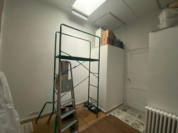 A small room mid-renovation, cluttered with a green scaffold, ladder, and painting supplies, suggesting a work-in-progress atmosphere. This image captures the essence of transformation, depicting a compact room undergoing renovation. The space is filled with the tools of change: a vibrant green scaffold stands at the center, accompanied by a ladder and painting essentials. Boxes and protective coverings hint at the room's transitory phase, awaiting a fresh coat of paint and new decor. The slanted ceiling suggests the room’s attic location, offering a cozy nook with potential for a charming redesign. The process captured here speaks to the room’s untapped possibilities, soon to be realized through the renovation journey.