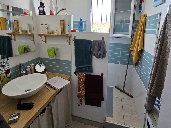 A well-organized bathroom with an above-counter basin on a wooden countertop, surrounded by personal care products. A shower area with blue tiles is visible, and various colored towels are hung neatly around the space. This photograph showcases a snug bathroom space that balances functionality and aesthetics. The above-counter white basin sits atop a wooden countertop, with daily toiletries arranged on shelves above, emphasizing an organized and accessible setup. The refreshing blue-tiled shower area adds a splash of color and a modern touch. The mix of green, blue, and yellow towels hanging around provides a vibrant contrast, while the natural light from the window enhances the room's welcoming atmosphere. Such an arrangement illustrates how even compact spaces can be transformed into inviting and practical areas.